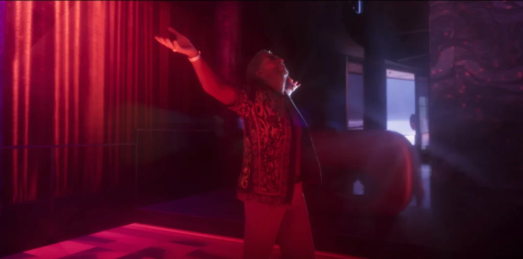 A male performer raises one arm while holding a microphone under red lighting in the GTA 6 launch trailer.